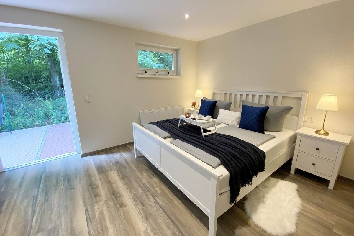Un dormitorio moderno con una cama blanca, dos mesitas de noche, un ventanal pequeño y vistas al exterior con vegetación.