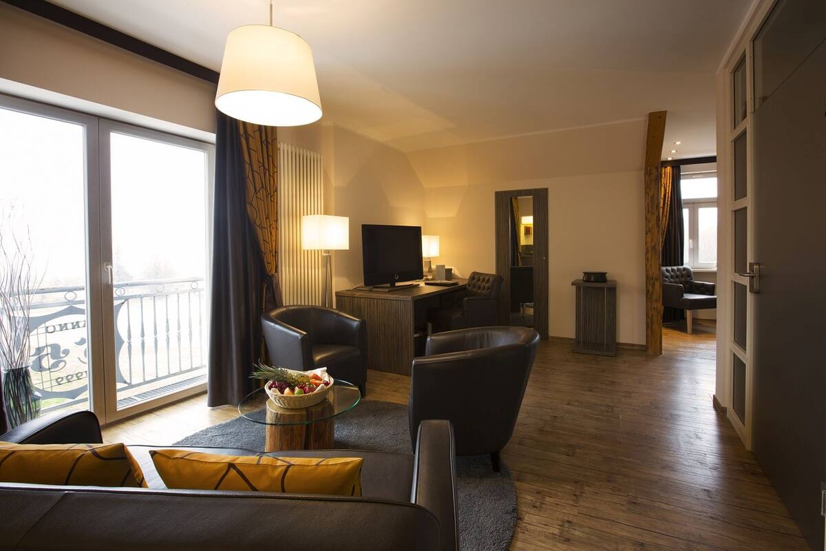 Una sala de estar moderna con sofá, sillones, una mesa de centro y televisor. Hay un balcón con barandilla y vistas al exterior.