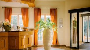 A reception area with a wooden counter, a large vase with flowers, and a view of the outdoors through large windows with orange curtains. - Regiohotel Berghotel Ilsenburg (Ilsenburg)