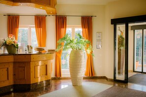 A reception area with a wooden counter, a large vase with flowers, and a view of the outdoors through large windows with orange curtains. - Regiohotel Berghotel Ilsenburg (Ilsenburg)