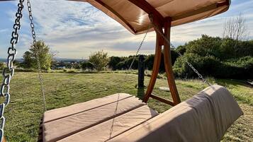 A wooden swing with a canopy, set in a grassy outdoor area with a view of the sky and distant buildings.