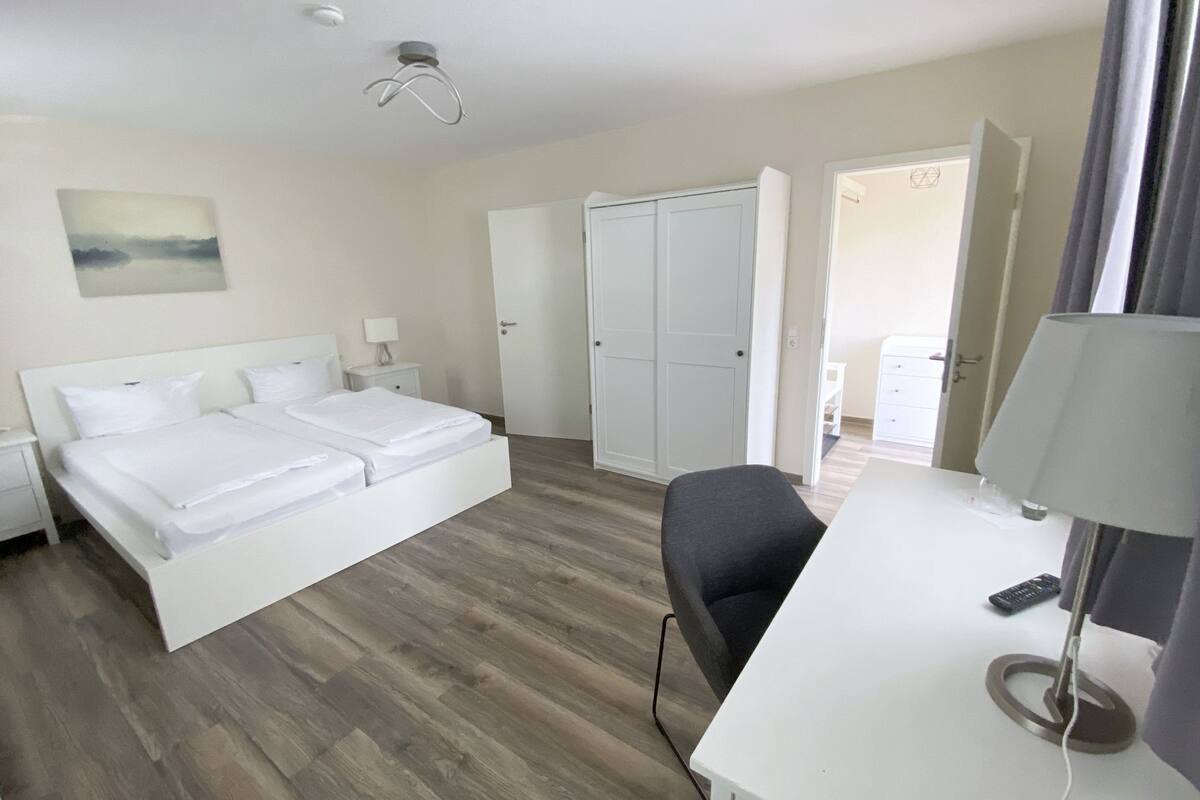 Una habitación de hotel con cama, escritorio, silla, lámpara de escritorio, control remoto y una puerta que da a otra habitación.