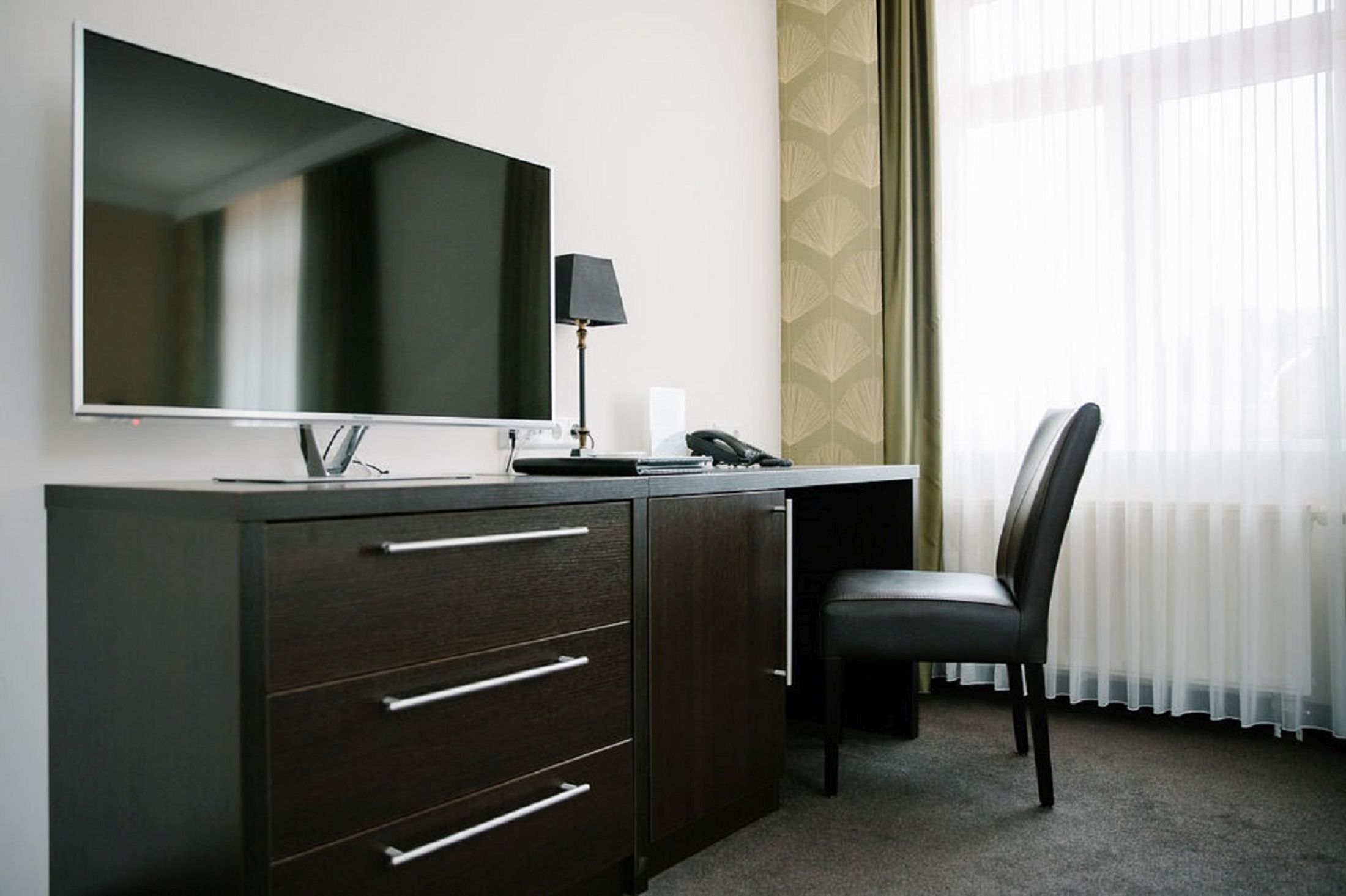 A hotel room with a large flat-screen TV mounted on a dark wooden cabinet, a desk with a lamp, a chair, and a telephone.
