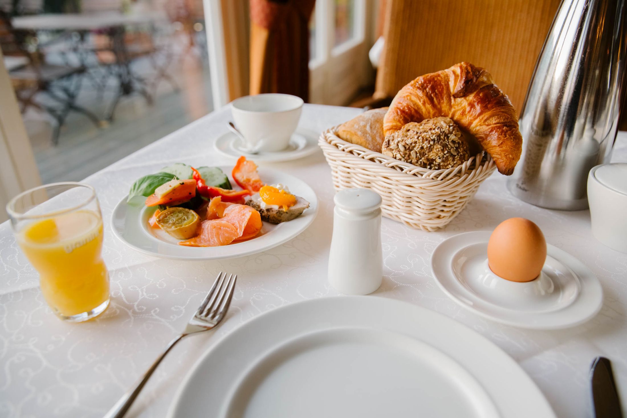 A breakfast setting with a croissant, bread basket, plate of fruit, egg, salt and pepper shakers, and a glass of orange juice on a table.