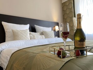 A neatly made bed with white linens and decorative pillows, a bedside table with a bottle of wine and a glass, and a tray with strawberries.