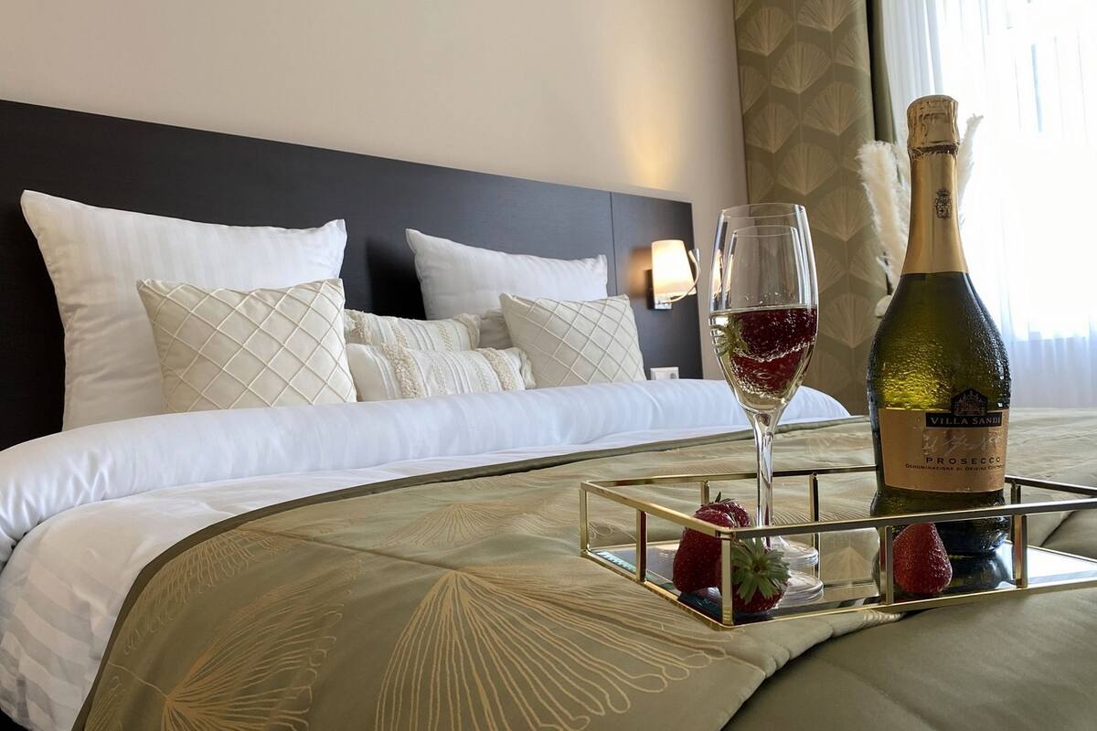 Una cama bien hecha con sábanas blancas y almohadillas decorativas, una mesita de noche con una botella de vino y una copa, y una bandeja con fresas.