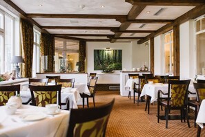 A dining area with tables set for service, a large forest-themed painting on the wall, and a view of the kitchen through a glass partition. - Regiohotel Berghotel Ilsenburg (Ilsenburg)