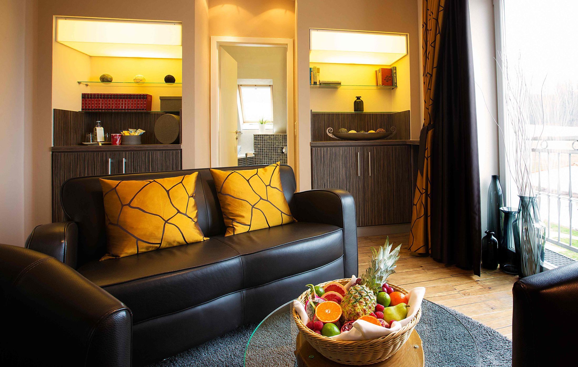 Ein modernes Wohnzimmer mit einem schwarzen Ledersofa, einer Obstschale auf einem Couchtisch und einem durch einen offenen Durchgang einsehbarer Küchenbereich.