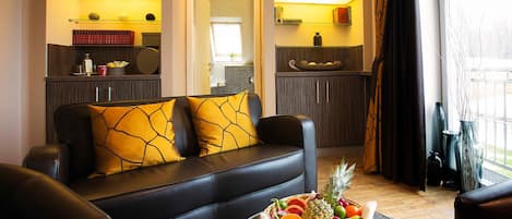 Uma sala de estar moderna com um sofá de couro preto, uma cesta de frutas em uma mesa de centro e uma área de cozinha visível por uma porta aberta.