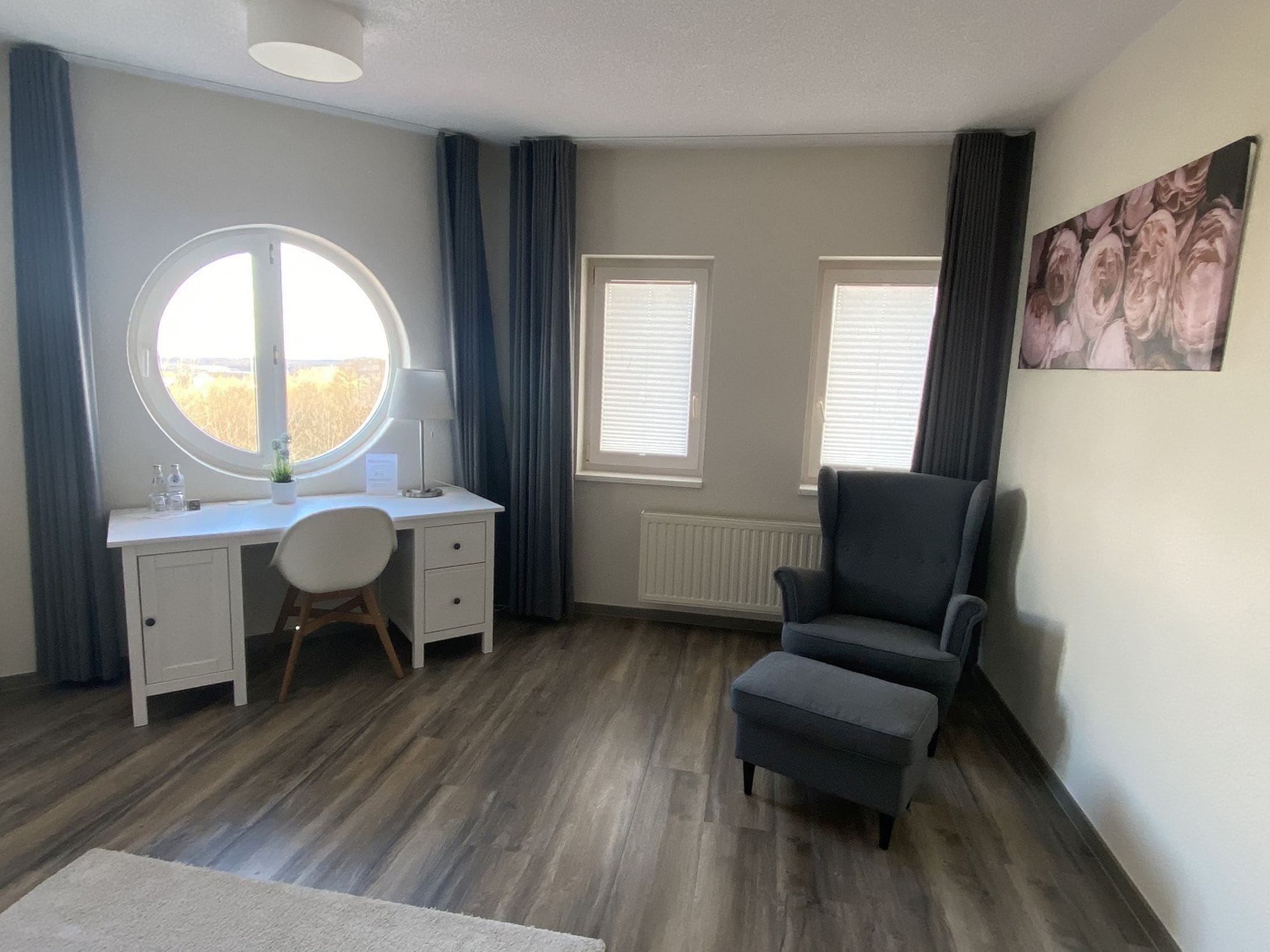 A room with a desk, chair, and ottoman, a round window, and a radiator.