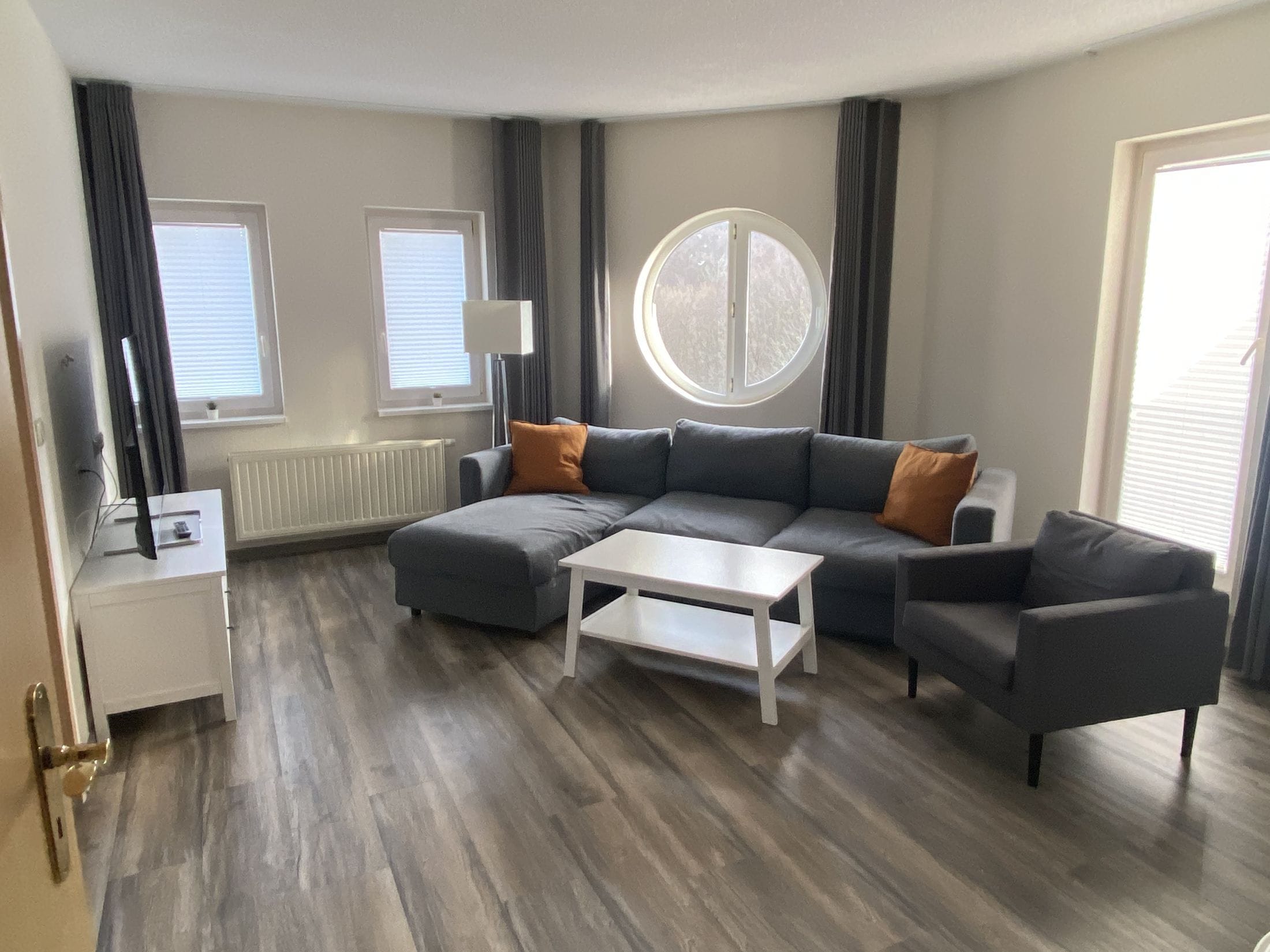 A modern living room with a sectional sofa, a coffee table, and a TV on a stand. There are two windows with blinds, a radiator, and a round window.