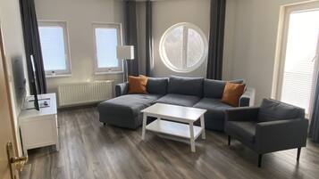 Ein modernes Wohnzimmer mit Ecksofa, Couchtisch und Fernseher auf einem Standfuß. Zwei Fenster mit Jalousien, ein Heizkörper und ein rundes Fenster sind zu sehen.