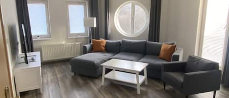 A modern living room with a sectional sofa, a coffee table, and a TV on a stand. There are two windows with blinds, a radiator, and a round window.