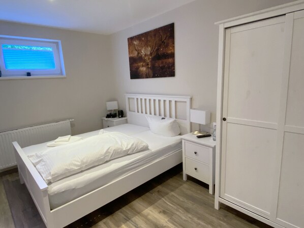 Un dormitorio con cama, mesitas de noche, armario, ventana con persianas y una obra de arte colgada en la pared.