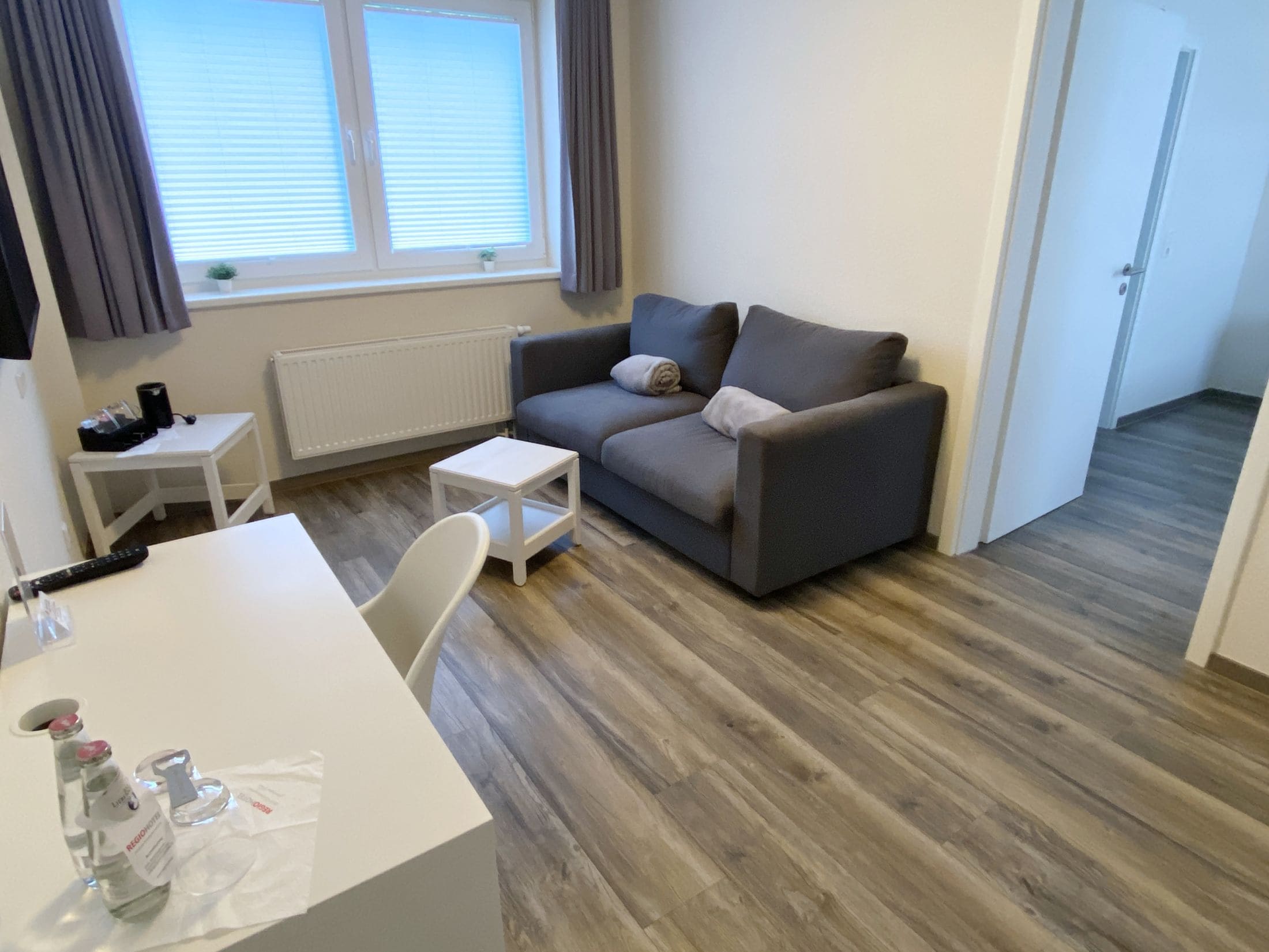 Un salon moderne avec un canapé gris, une petite table blanche et une chaise blanche. Il y a une fenêtre avec des stores et un radiateur au mur.