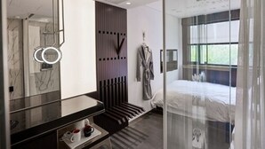 A modern hotel room with a large mirror, a bedside table with a cup and saucer, a bed with a white duvet, and a window with curtains.
