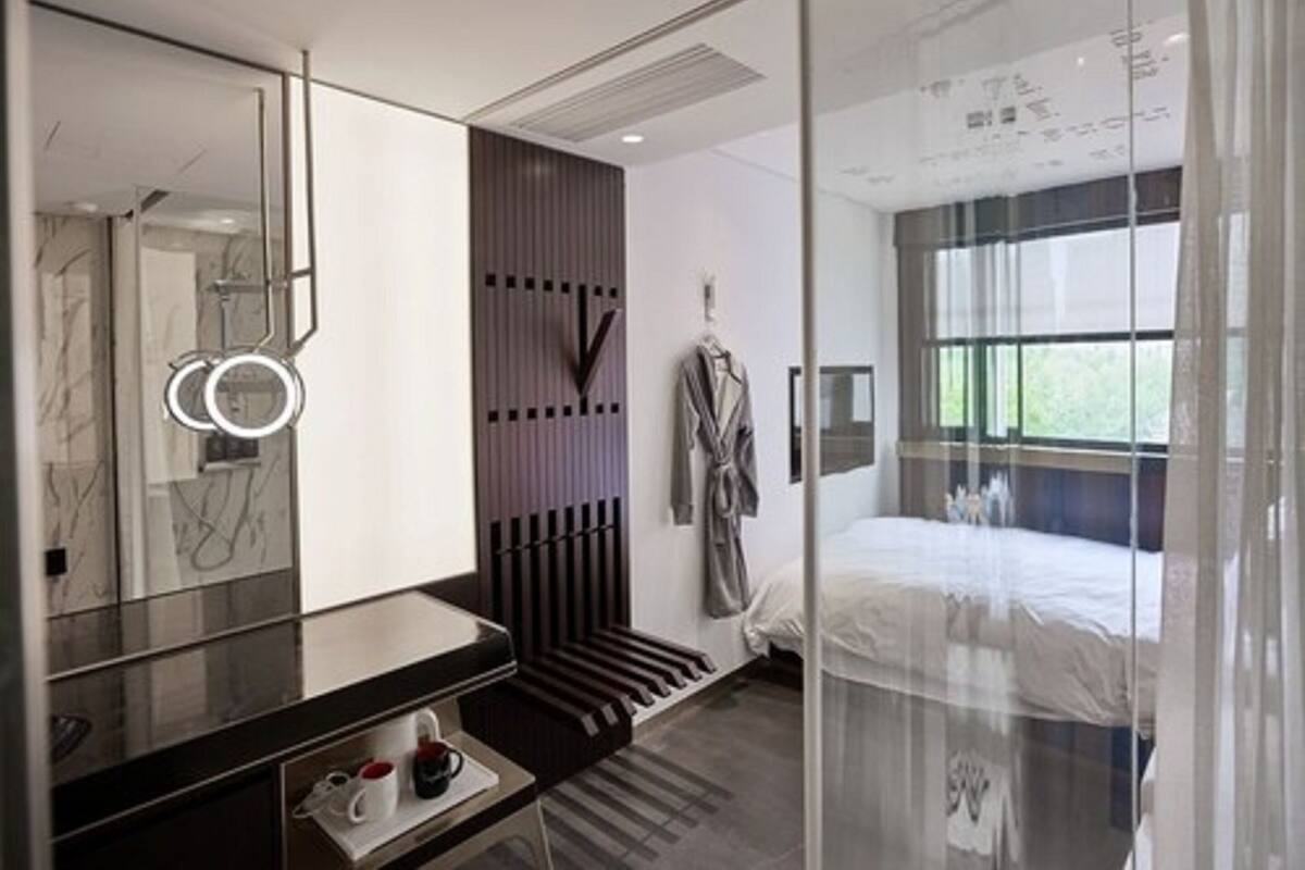 Een moderne hotelkamer met een grote spiegel, een nachtkastje met kop en schotel, een bed met witte dekbedovertrek en een raam met gordijnen.