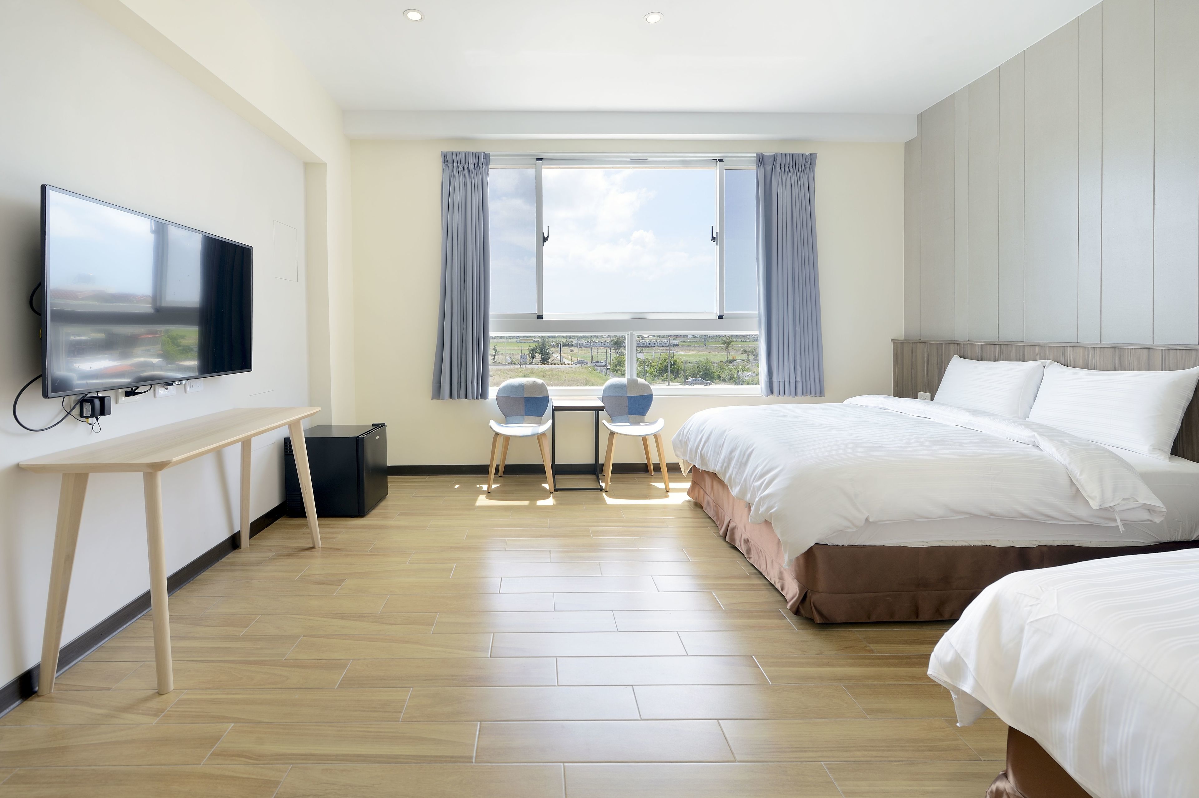 A hotel room with a bed, a TV, a desk, two chairs, and a window with curtains.