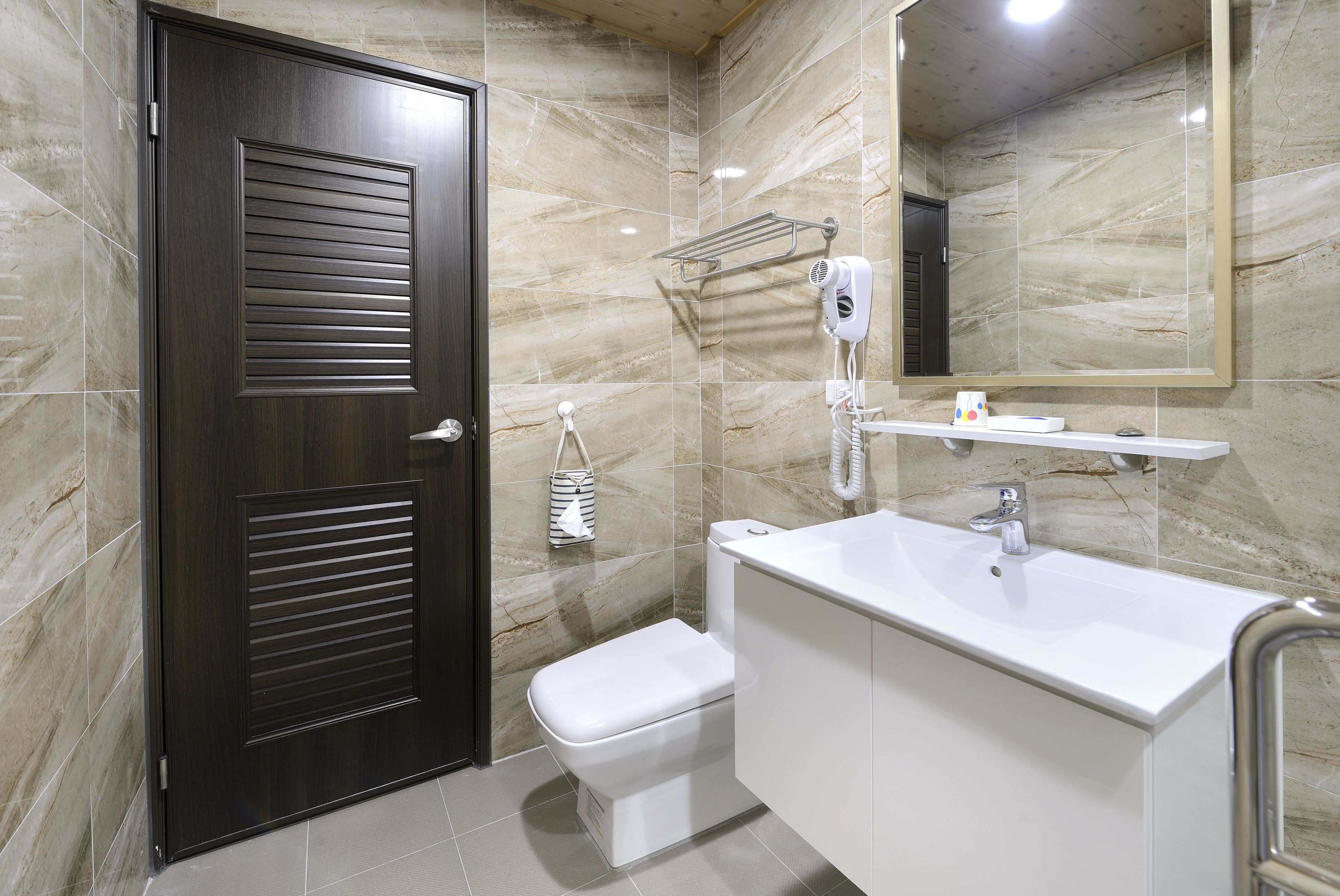 Une salle de bain moderne avec une porte en bois foncé, des toilettes blanches, un lavabo blanc avec un miroir au-dessus et un porte-serviettes muni d’un sèche-cheveux.