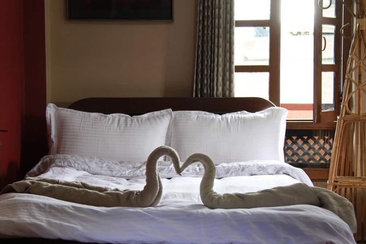 A hotel room with a bed, white linens, a heart-shaped towel arrangement, a window with curtains, and a wicker chair.