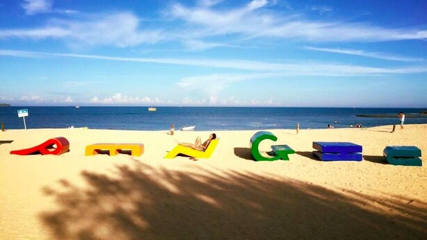 Une plage avec un ciel dégagé, quelques bateaux sur l’eau et des lettres colorées dessinées dans le sable.
