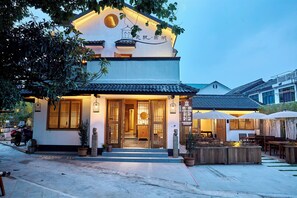Un bâtiment de style japonais traditionnel, avec un toit recouvert de tuiles, une porte d’entrée en bois et un petit jardin agrémenté d’un espace pour s’asseoir.