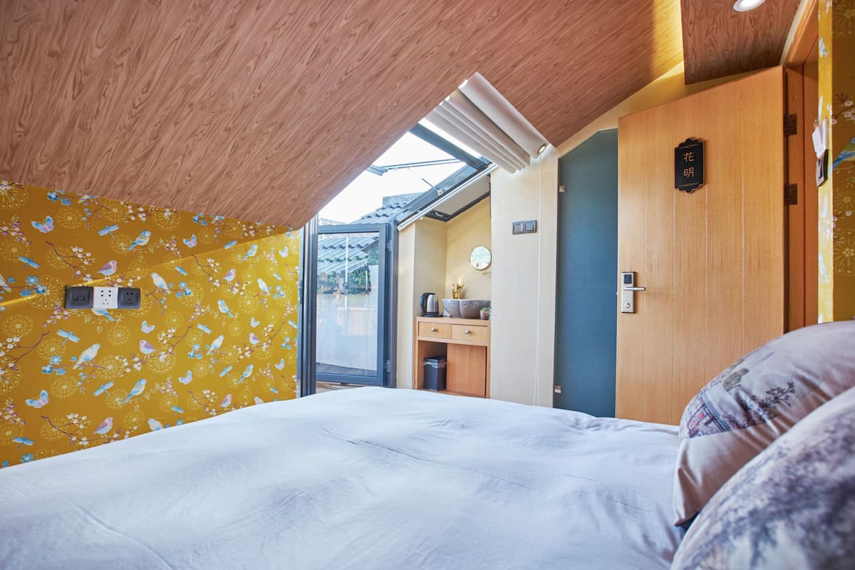 Een slaapkamer met dakraam, houten plafond, bed, een klein keukenhoekje en een deur met een bordje.