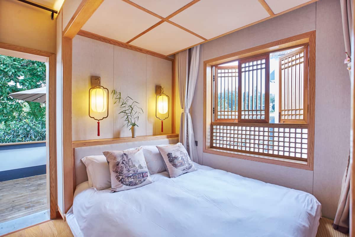 Een kamer in traditionele stijl met een bed, decoratieve wandlampen, een raam met houten spijlen en uitzicht op groen buiten.