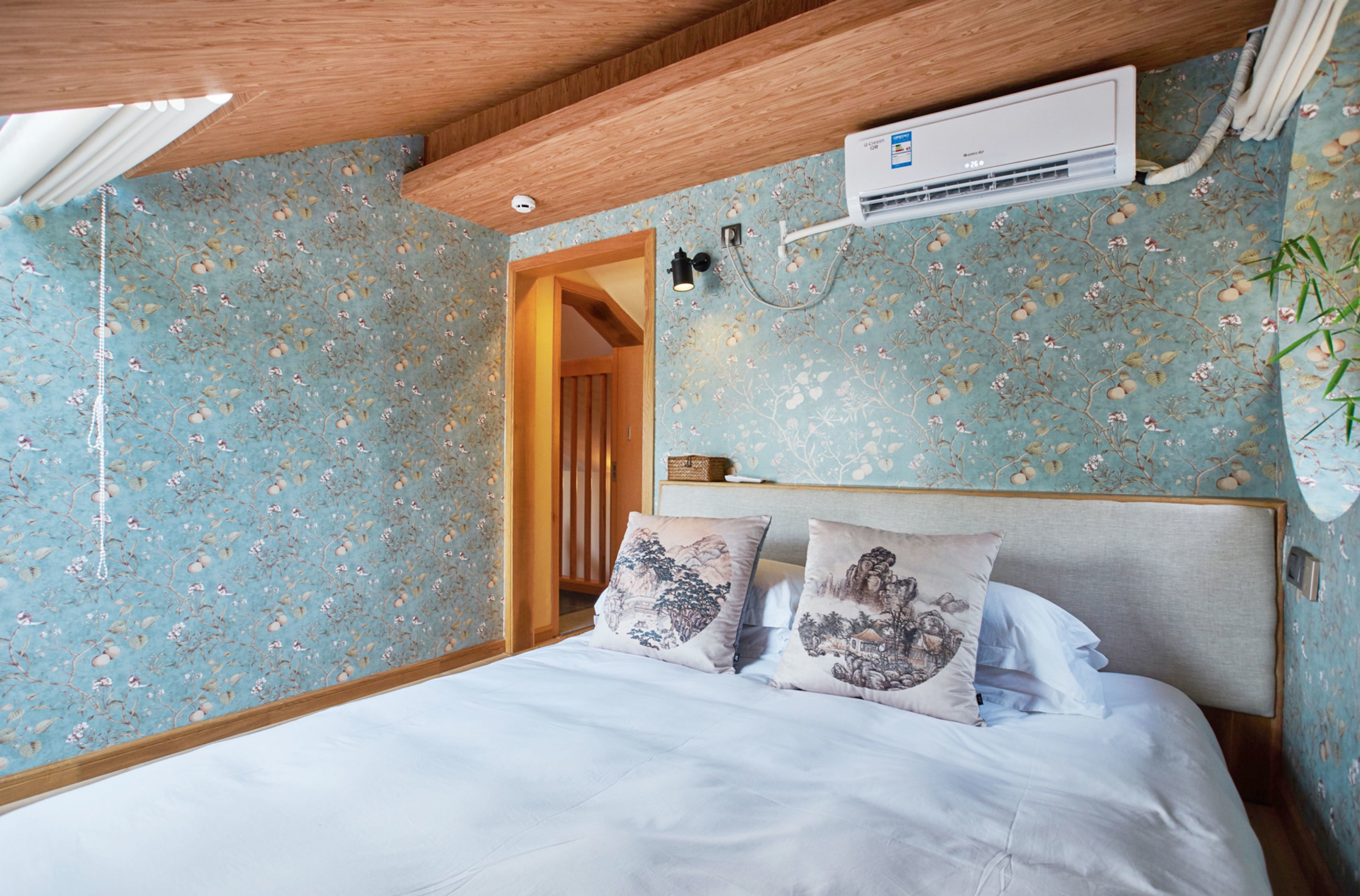 A bedroom with floral wallpaper, a wooden ceiling, a bed with white bedding, two decorative pillows, and an air conditioning unit.