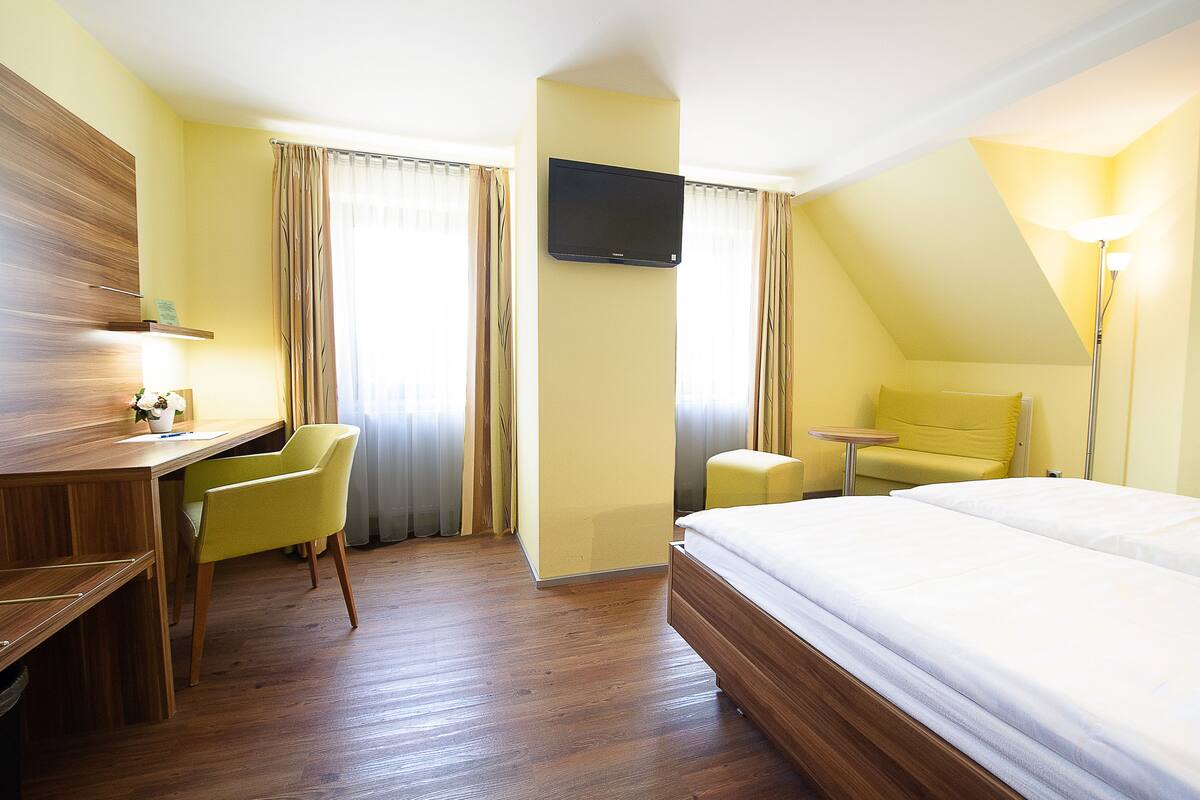 A hotel room with a bed, a desk with a chair, a TV mounted on the wall, and a lamp.