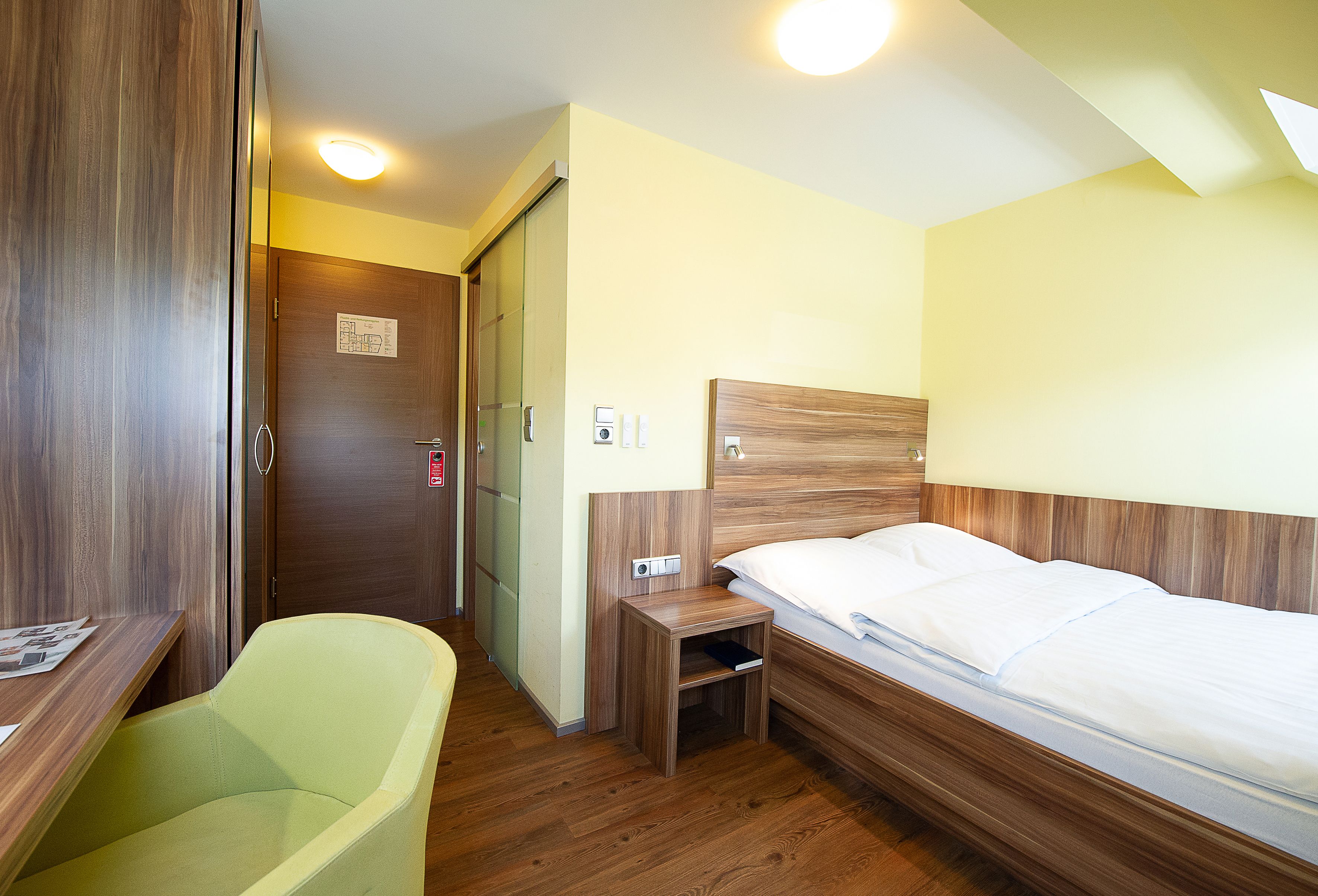 A compact hotel room with a bed, a small bedside table, a chair, and a wardrobe.
