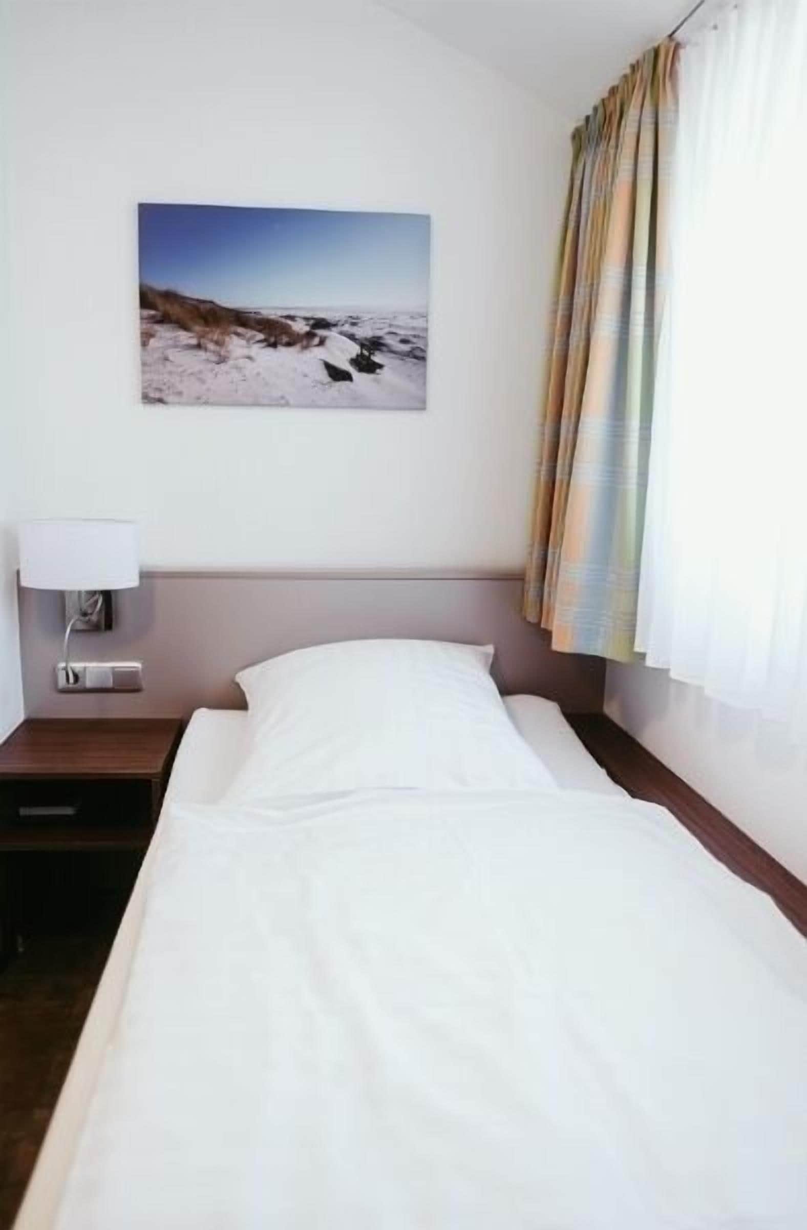 A hotel room with a bed, a nightstand, a lamp, a painting of a snowy landscape, and a curtain.