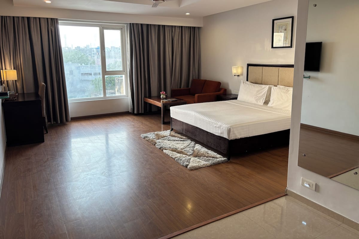 A modern hotel room with a large bed, a desk with a lamp, a chair, a TV, and a view of the city through the window.