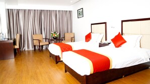 A hotel room with two beds, a wooden floor, a desk with a chair, and a large window with curtains.