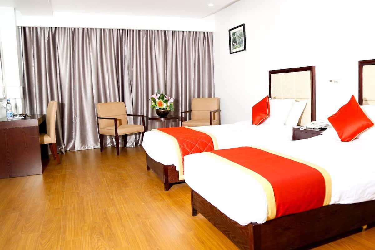 A hotel room with two beds, a wooden floor, a desk with a chair, and a large window with curtains.