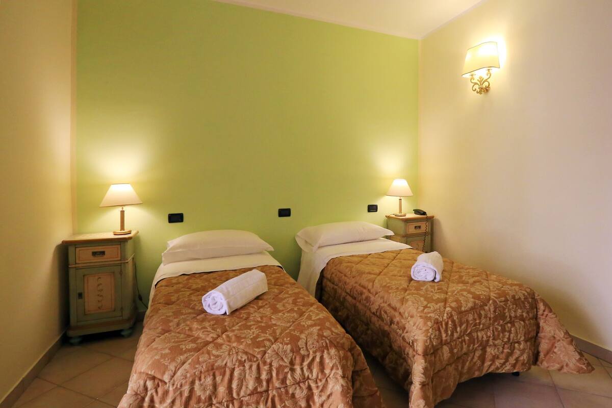 Ein Hotelzimmer mit zwei Betten, jeweils mit einem blumigem Überwurf und einem zusammengerollten Handtuch. An jedem Bett befinden sich Nachttischlampen und ein Nachttisch mit einem Telefon.