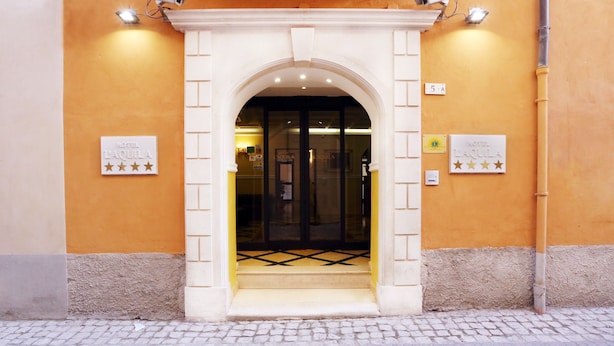 The entrance to a hotel with a white archway, a glass door, and a tiled floor. There are signs on the wall indicating the hotel's name and a star rating.