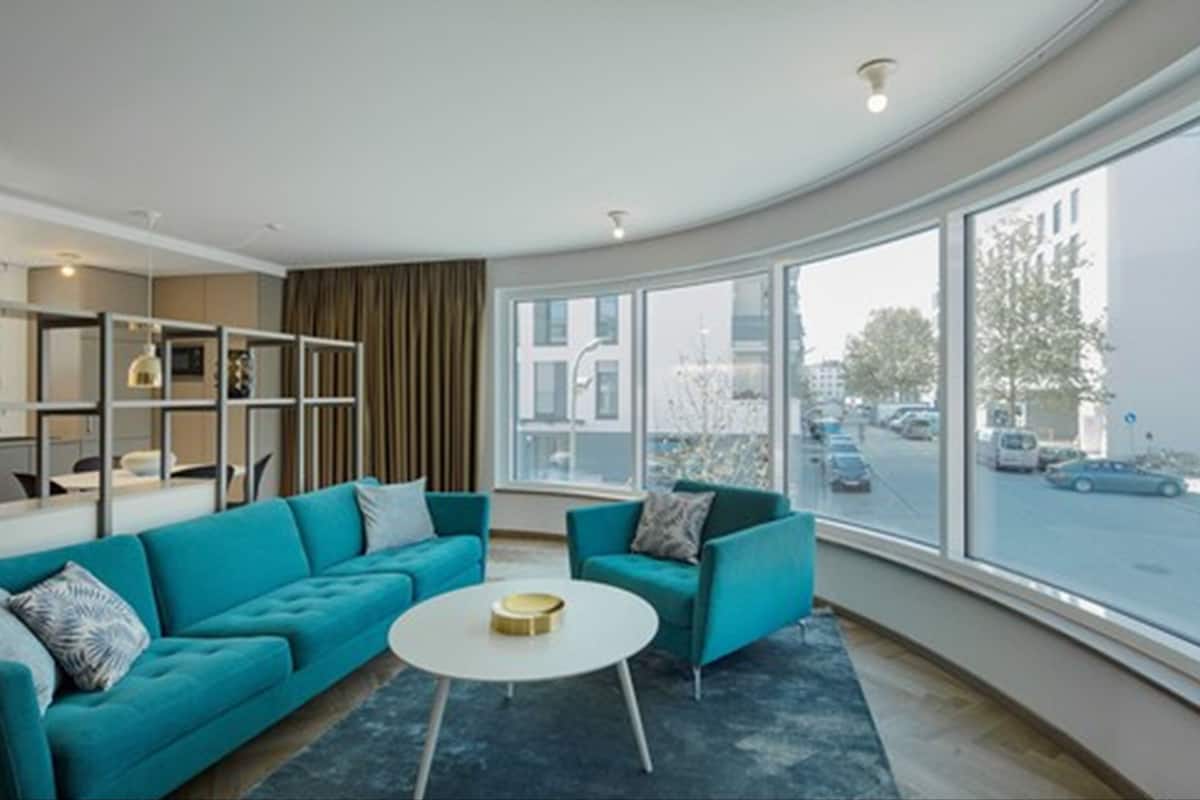 A modern living room with teal sofas, a white coffee table, and large windows offering a city view.