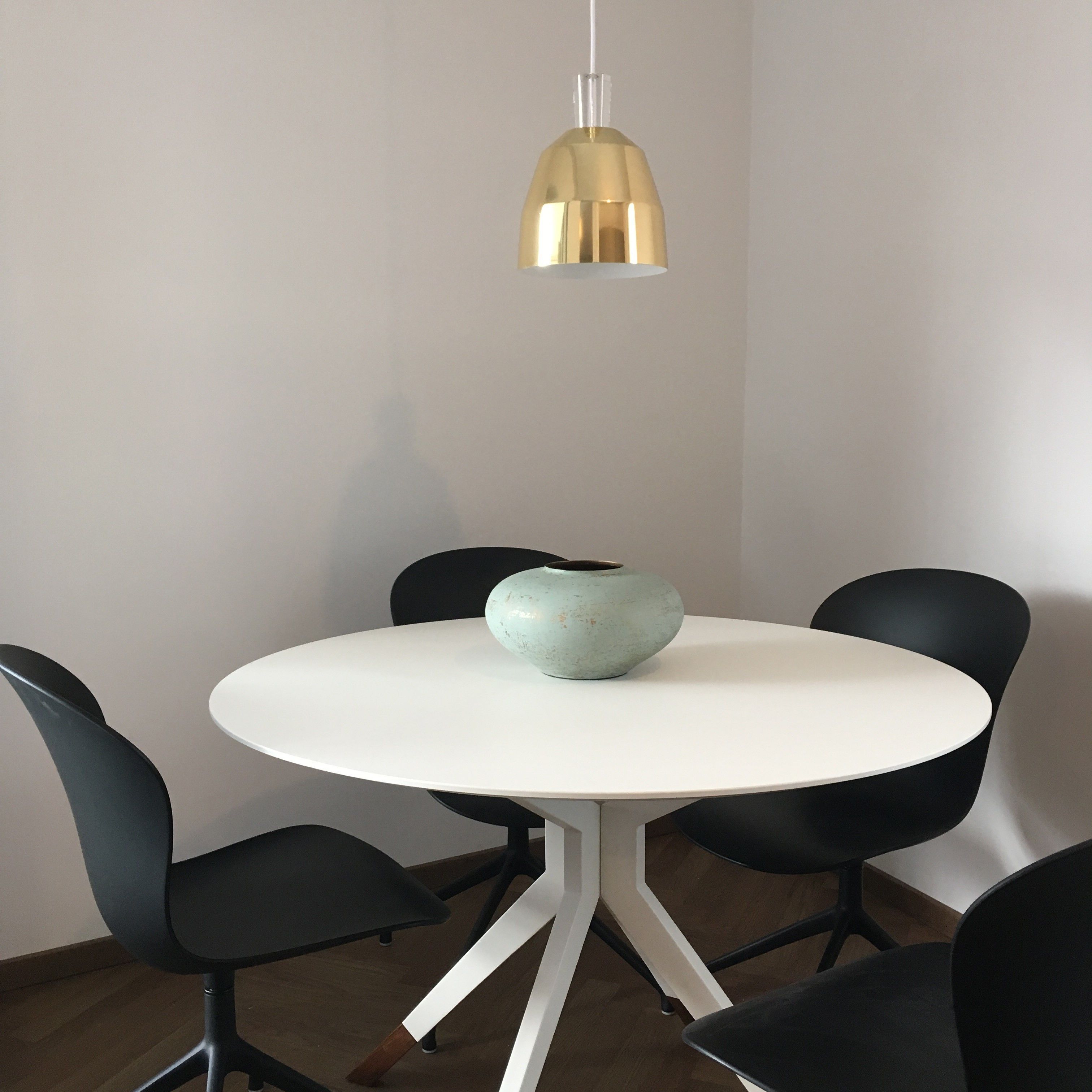 A round white table with a light blue vase, surrounded by black chairs. A gold pendant light hangs above.