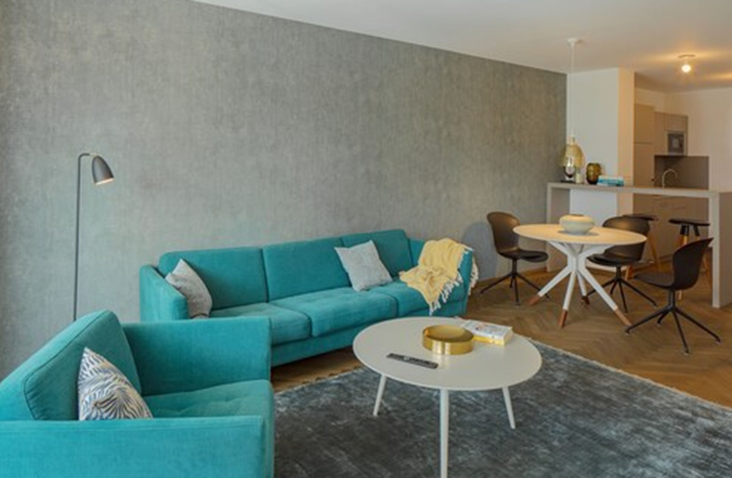 Ein modernes Wohnzimmer mit einem türkisblauen Sofa, einem runden Couchtisch und einem Essbereich mit weißem Tisch und Stühlen.