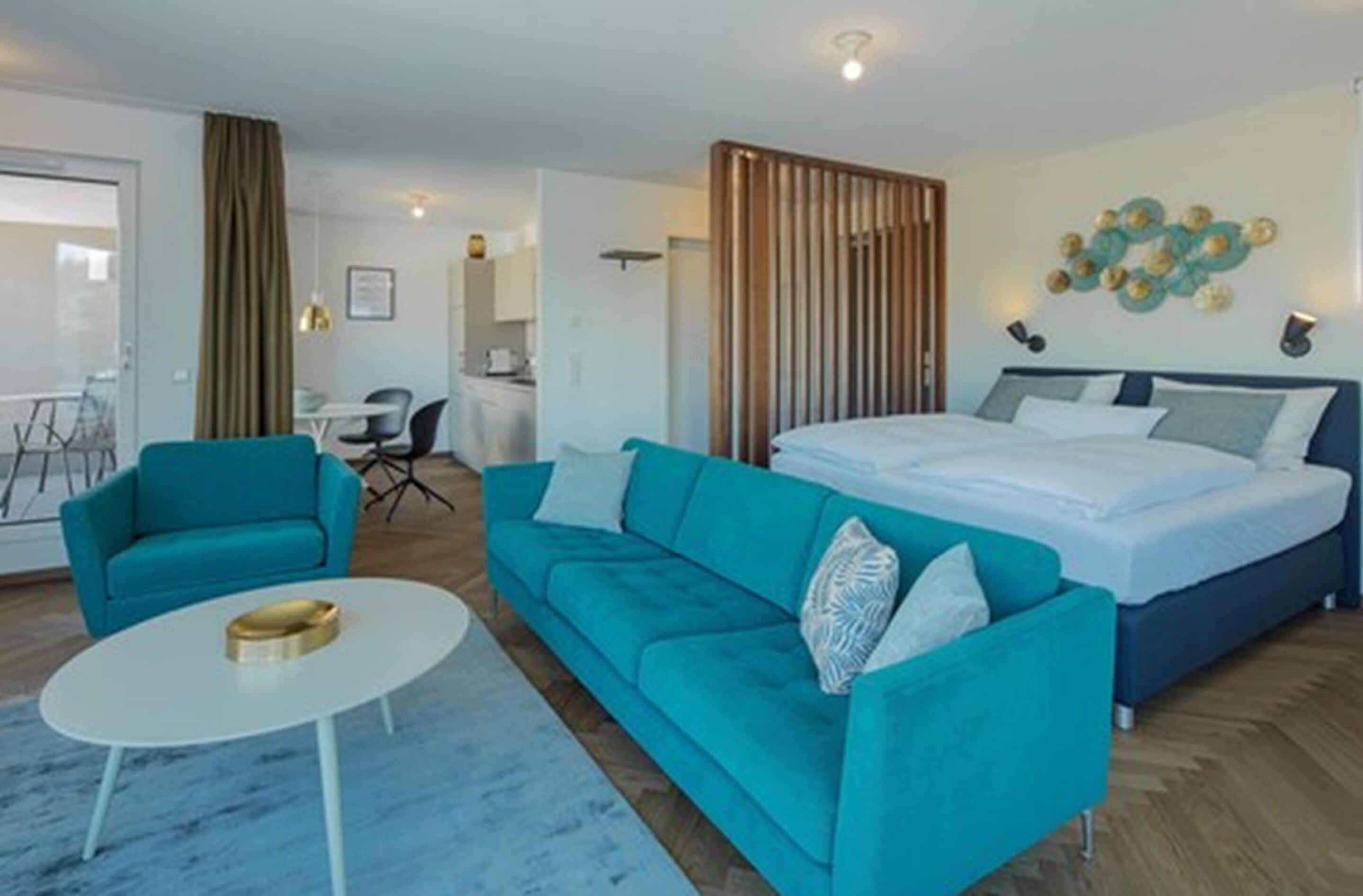 A modern hotel room with a bed, two teal sofas, a white coffee table, and a wooden partition.