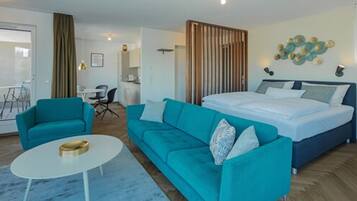 A modern hotel room with a bed, two teal sofas, a white coffee table, and a wooden partition.