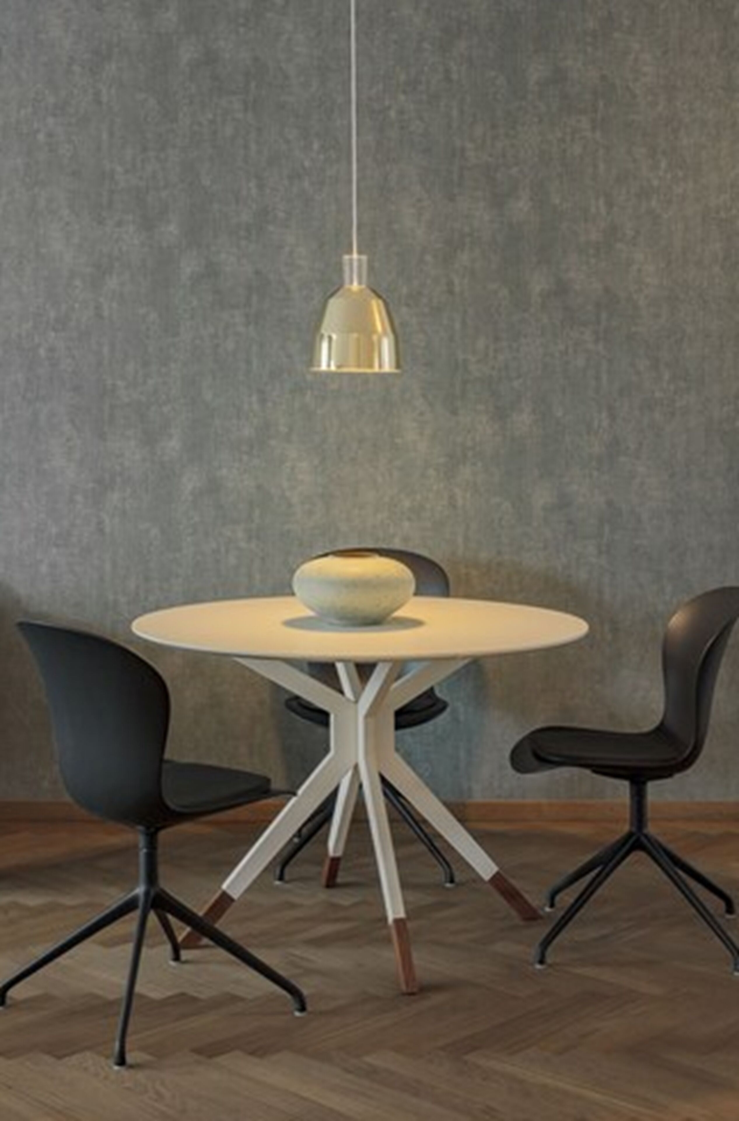 Une table de salle à manger ronde, dotée d’un pied central blanc et surmontée d’un grand bol. Deux chaises noires à l’assise originale, avec un pied tripode, sont placées de part et d’autre de la table. Un luminaire suspendu éclaire l’ensemble.