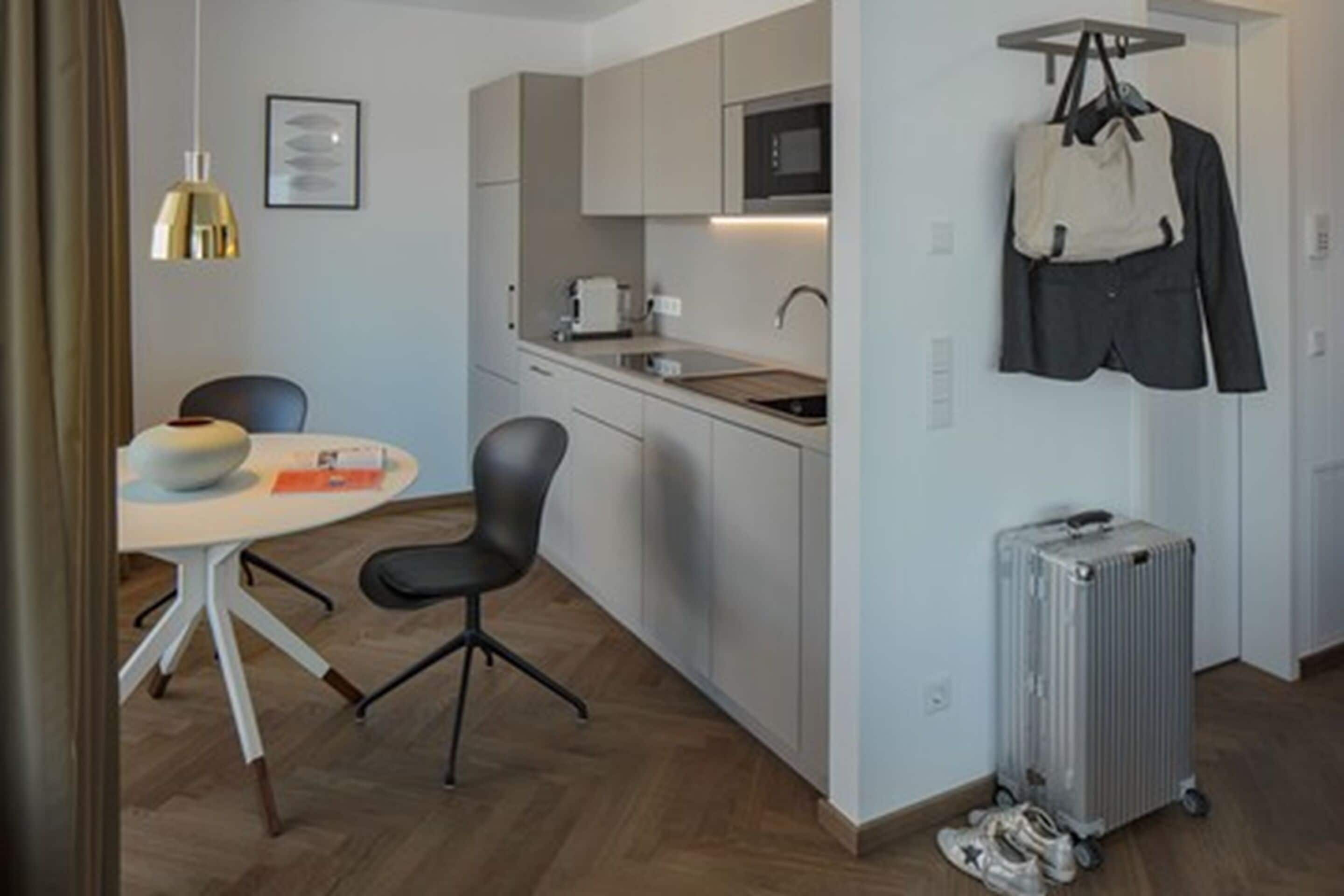 A compact kitchen with a microwave, a small sink, and a chair. There is a table with a bowl and a book. A suitcase and a bag are hanging on a rack.