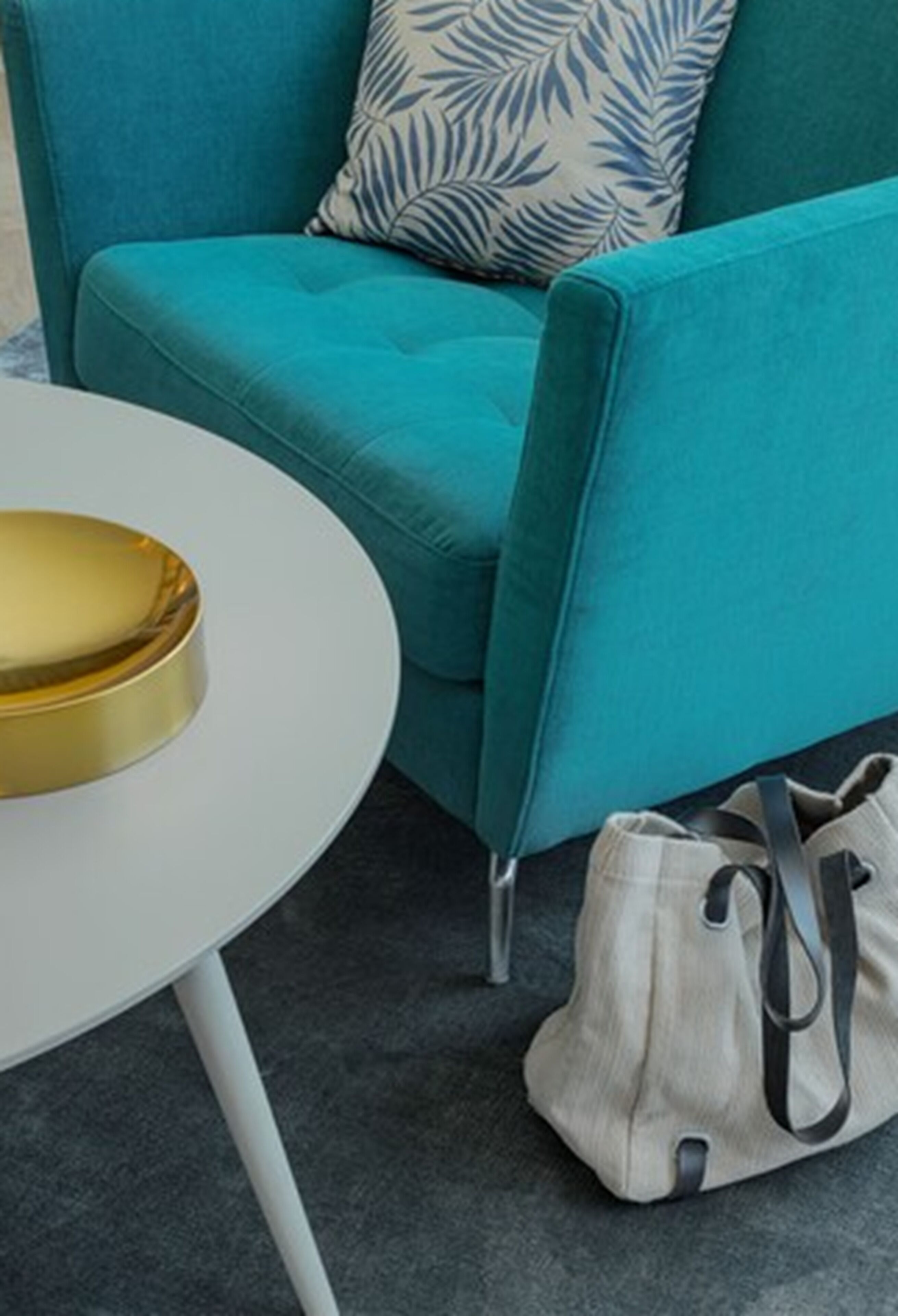 A teal sofa with a leaf-patterned pillow, a white table with a gold tray, and a beige bag on the floor.