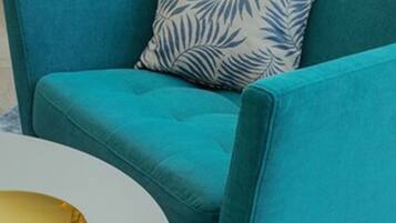 A teal sofa with a leaf-patterned pillow, a white table with a gold tray, and a beige bag on the floor.