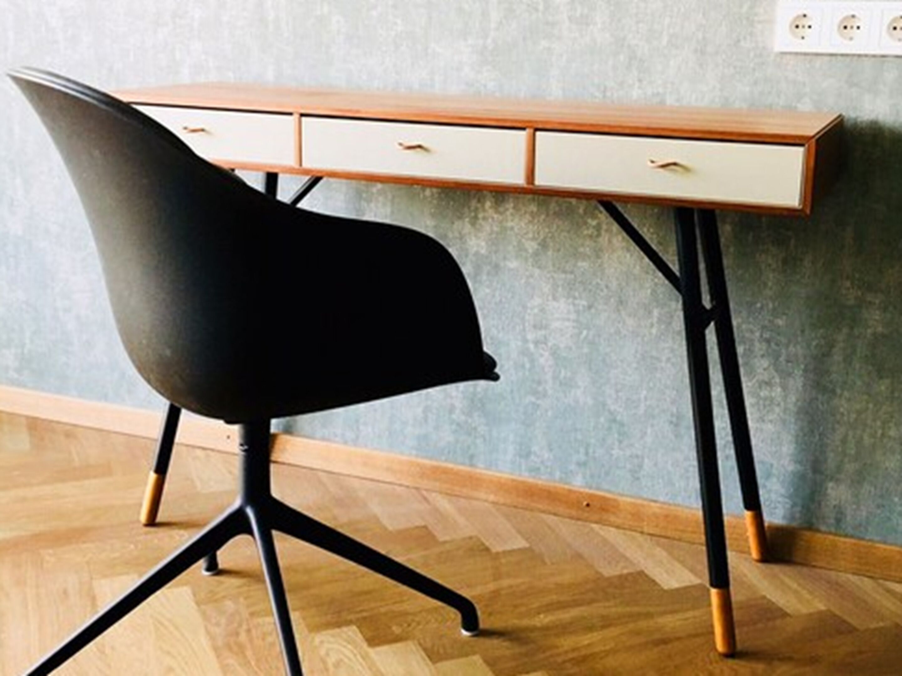 Un escritorio moderno con una silla negra, patas de madera y superficie clara.