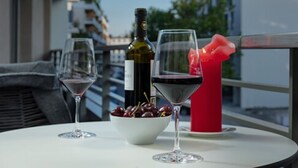 A table with two wine glasses, a bottle of wine, a bowl of cherries, and a red candle on a balcony with a railing and buildings in the background. - DD Suites Serviced Apartments (Munich)