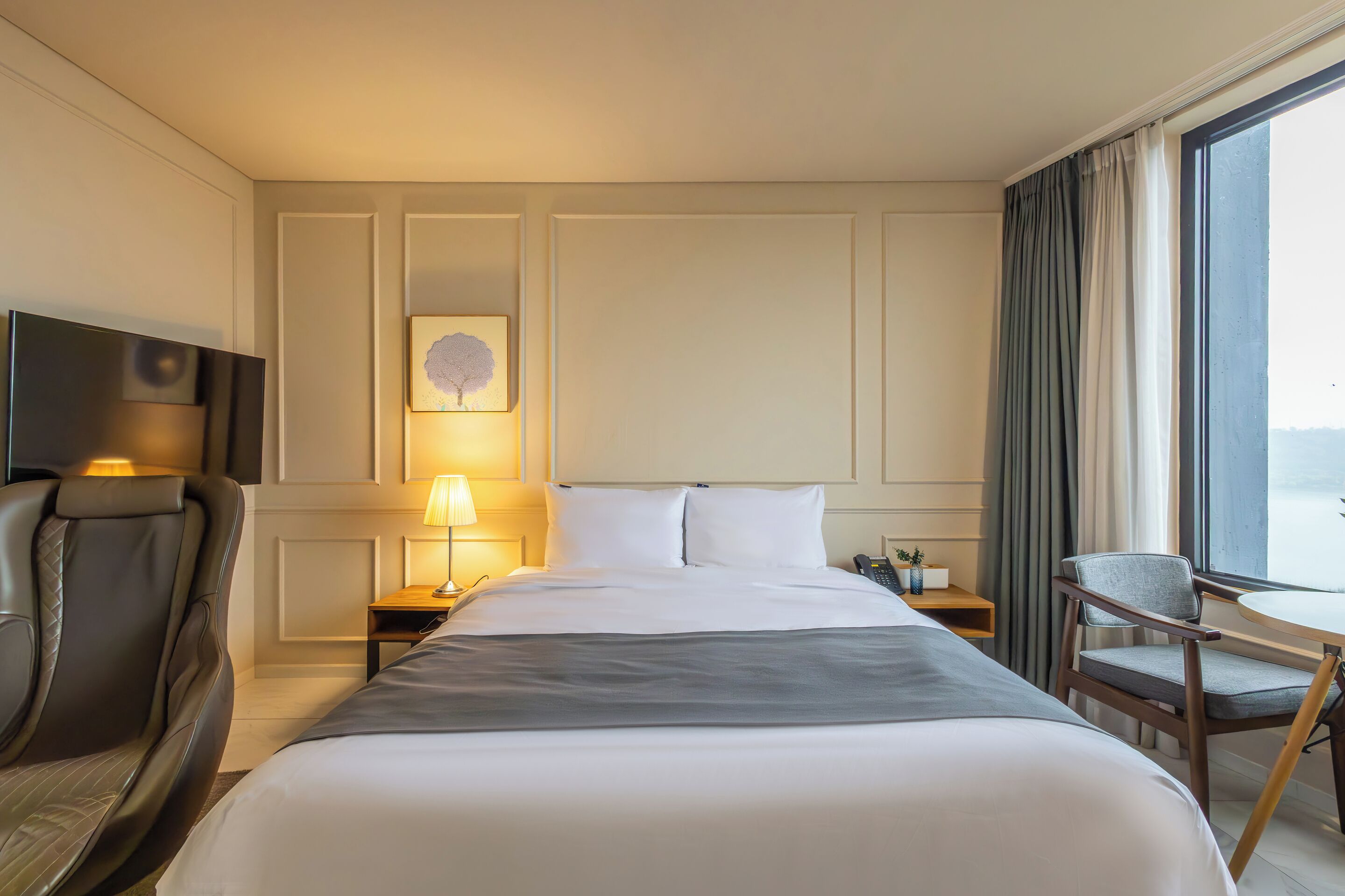A hotel room with a large bed, a desk with a lamp, a chair, and a TV.