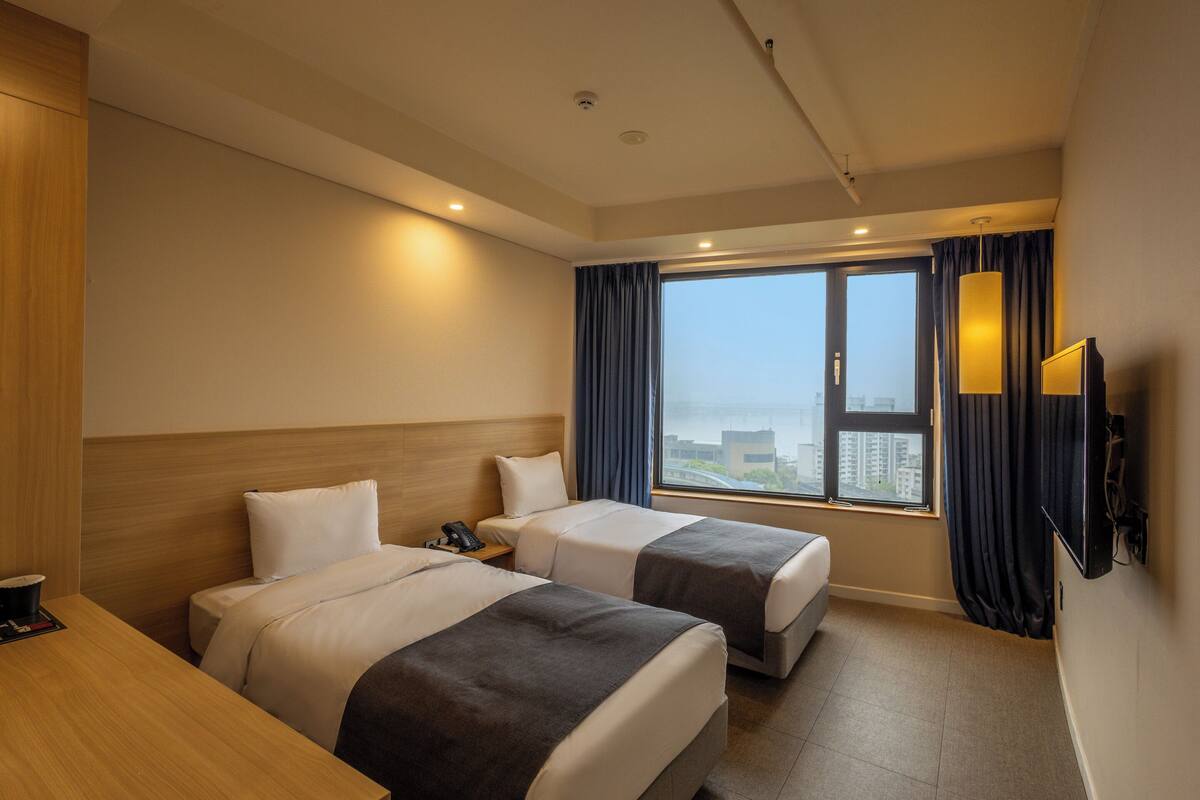 A hotel room with two beds, a desk, a phone, a TV, and a large window with curtains.
